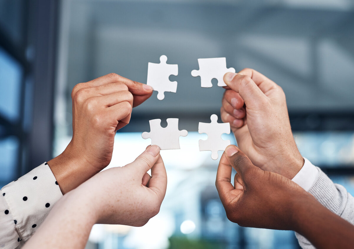 Multiple hands holding white puzzle pieces and bringing them together, symbolizing collaboration, partnership, and joint problem-solving.
