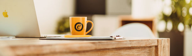 Nahaufnahme eines Schreibtisches mit einer orangenen Tasse, die das Logo der Trusted Shops GmbH aufgedruckt hat.