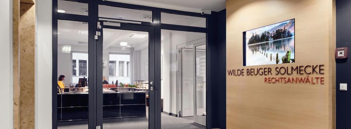 Entree van het Keulse advocatenkantoor WILDE BEUGER SOLMECKE, met een moderne receptieruimte voorzien van een glazen gevel. Op een met hout afgewerkte wand staat de naam 'WILDE BEUGER SOLMECKE RECHTSANW&Auml;LTE' weergegeven. Achter de glazen deuren zijn kantoorruimtes zichtbaar, wat de open en professionele uitstraling van het kantoor benadrukt.