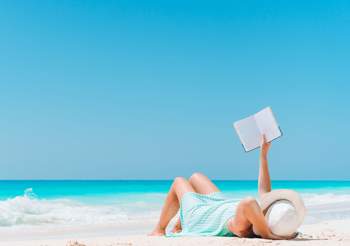 Person liest entspannt mit einem Buch am Strand vor t&uuml;rkisfarbenem Meer und blauem Himmel.