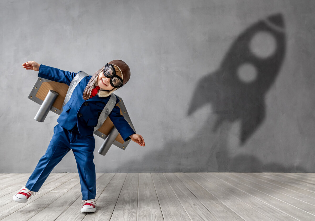 Child wearing a blue pilot costume with an aviator cap, goggles, and a homemade cardboard airplane. A rocket-shaped shadow is visible on the wall behind. The scene symbolizes vision, innovation, growth, and the courage to explore new possibilities.