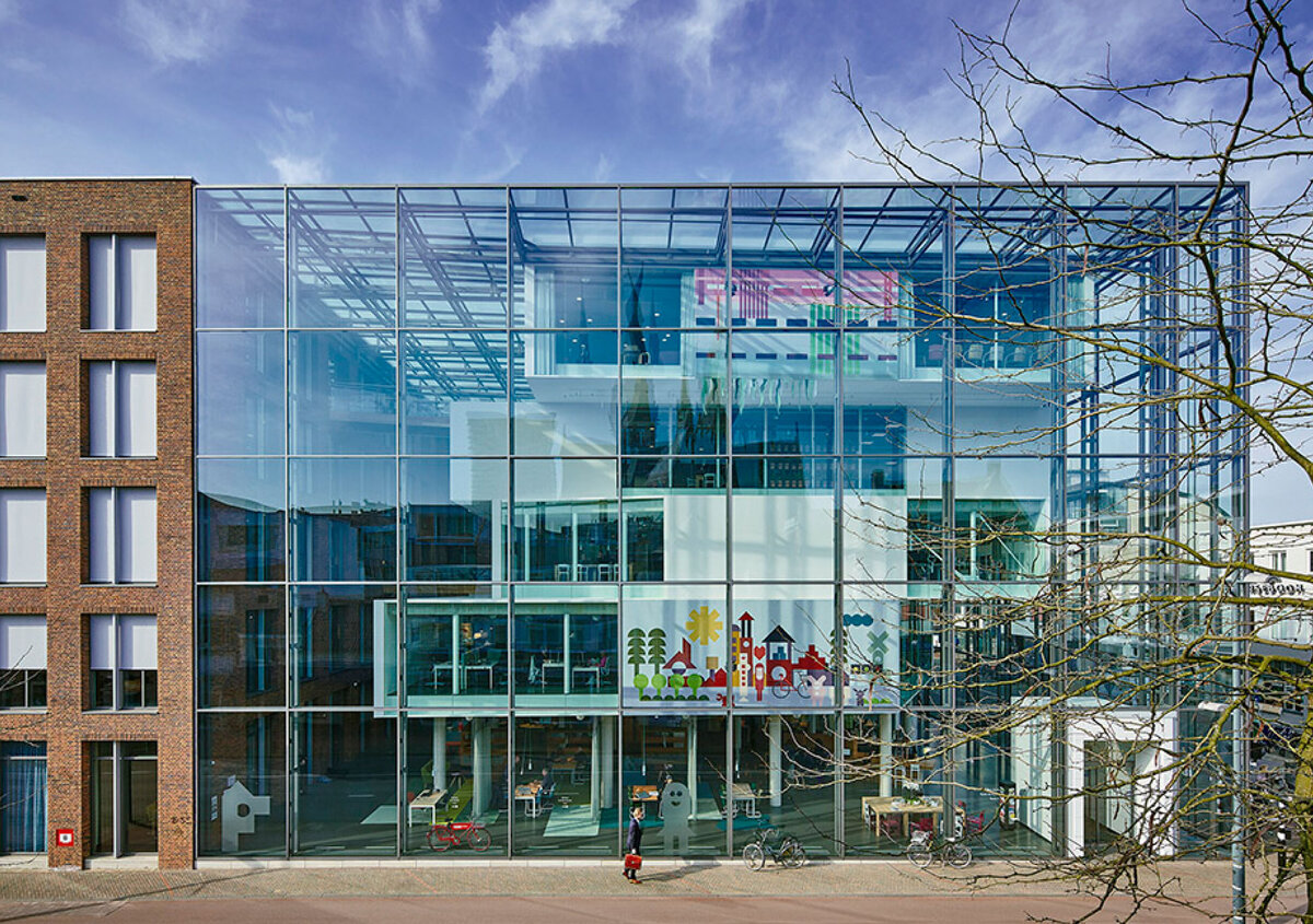 Modernes B&uuml;rogeb&auml;ude mit gro&szlig;er Glasfassade und mehreren sichtbaren Etagen. Durch die verglaste Front sind offene Arbeitsbereiche, Besprechungsr&auml;ume und farbige Gestaltungselemente erkennbar. Vor dem Geb&auml;ude verl&auml;uft ein Gehweg mit einigen Fahrr&auml;dern und einer Person. Rechts ragen die &Auml;ste eines kahlen Baumes ins Bild, dar&uuml;ber ein heller Himmel mit leichten Wolken.