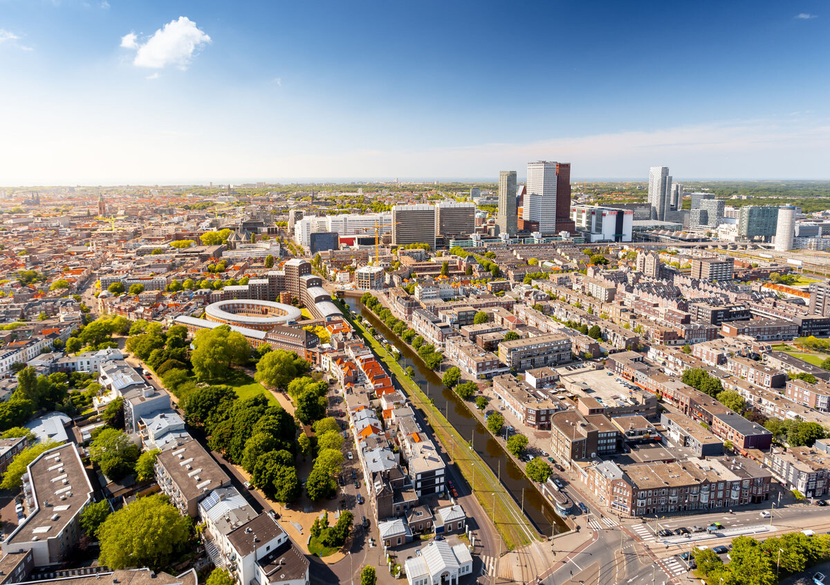 Flygvy &ouml;ver ett vidstr&auml;ckt stadslandskap en solig dag. I f&ouml;rgrunden syns bostadsbyggnader, tr&auml;dall&eacute;er och en rak kanal som l&ouml;per genom omr&aring;det. I bildens mitt ligger t&auml;tt bebyggda stadsdelar med ett karakteristiskt cirkul&auml;rt byggnadskomplex. I bakgrunden reser sig en modern stadssiluett med flera h&ouml;ghus och kontorsbyggnader. Ovanf&ouml;r staden breder en klarbl&aring; himmel ut sig med endast n&aring;gra f&aring; moln.