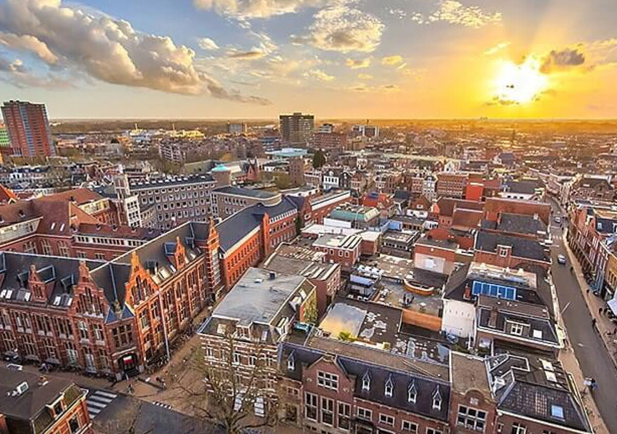Aerial view of a historic city center at sunset. In the foreground, densely packed residential blocks and distinctive red-brick buildings with ornate stepped gables shape the urban landscape. Numerous streets and narrow alleys wind between the buildings, while the city stretches toward the horizon. The low sun bathes the scene in warm, golden light, illuminating the clouds overhead. In the background, modern high-rise buildings and newer urban districts create a striking contrast to the historic architecture.