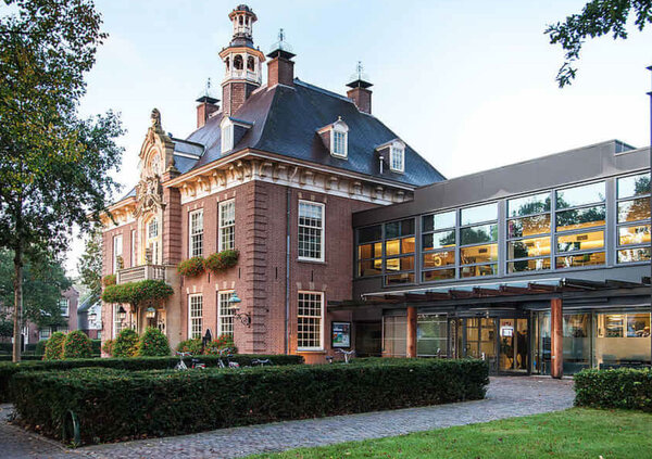 Historic brick building with distinguished architecture, a steep mansard roof, and a small tower. Attached to the right is a modern extension featuring a large glass fa&ccedil;ade and a glazed entrance area. Well-maintained hedges, lawns, and a paved walkway lead to the entrance. Several bicycles are parked in front of the building. Captured in daylight, the image highlights the successful combination of historic and contemporary architecture.