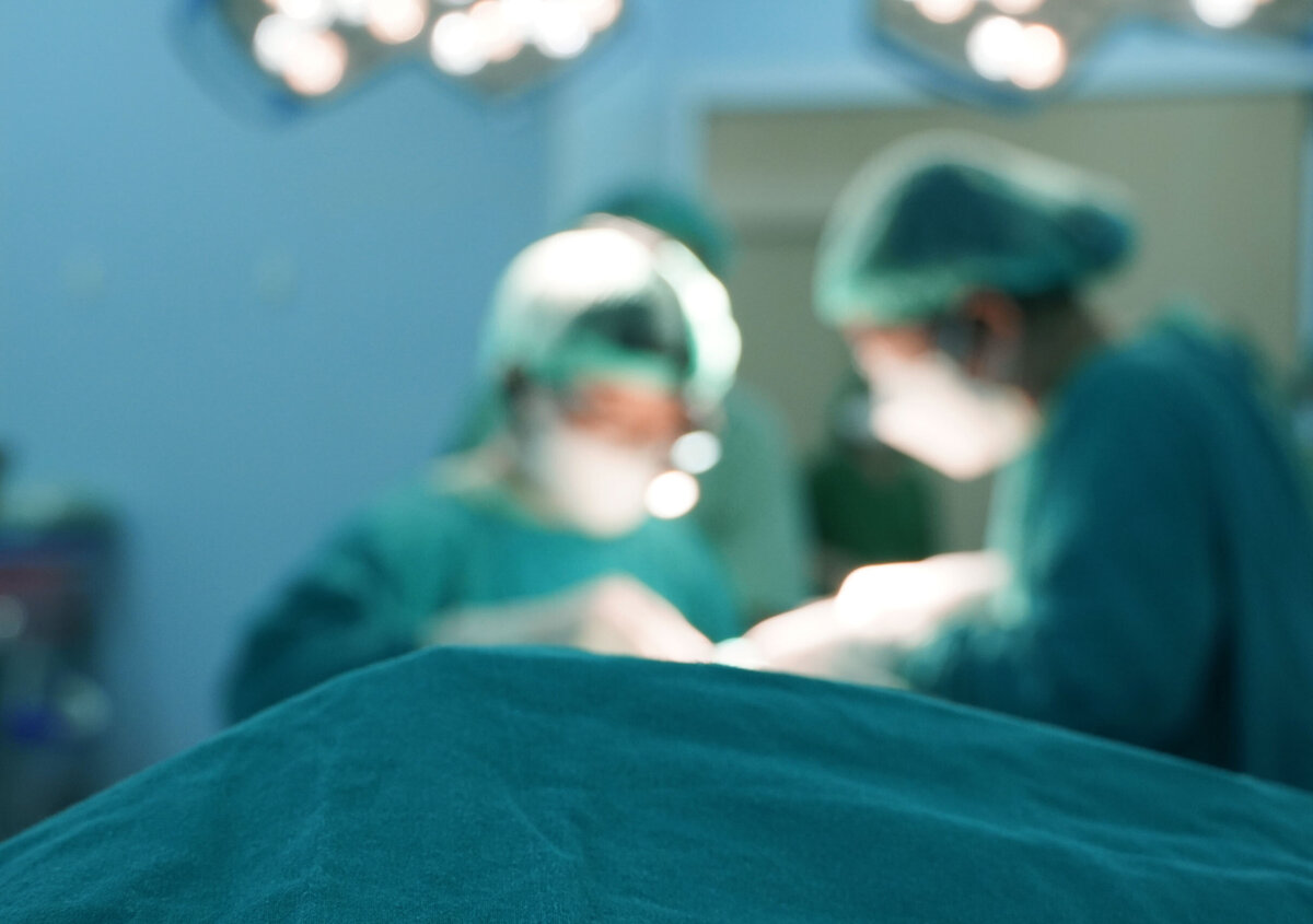 Blurred view of a surgical procedure in an operating room, with medical professionals in protective clothing working around a patient under bright surgical lights.