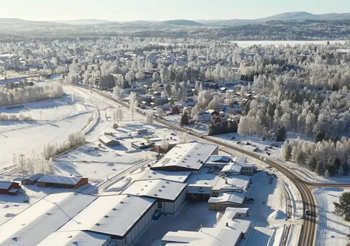 Aerial view of a snow-covered landscape in winter. In the foreground, there is a large industrial and office complex consisting of several warehouse and administrative buildings, with snow-covered roofs.