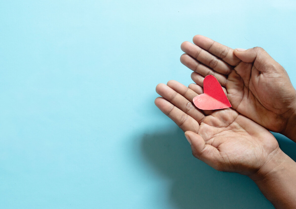 Two open hands gently hold a small red paper heart against a light blue background. The heart rests in the center of the palms and serves as the focal point of the image. The simple composition leaves ample empty space around the subject, while the soft colors convey themes of care, support, compassion, and social responsibility.