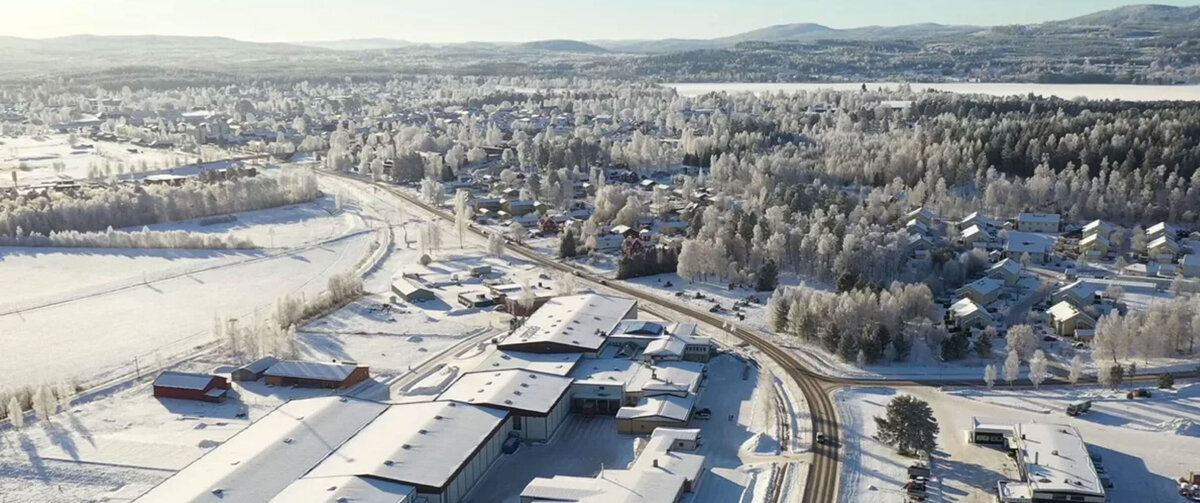 Aerial view of a snow-covered landscape in winter. In the foreground, there is a large industrial and office complex consisting of several warehouse and administrative buildings, with snow-covered roofs.