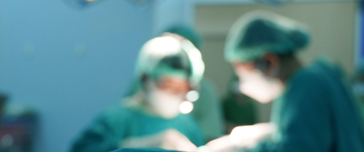 Suddig vy av ett kirurgiskt ingrepp i en operationssal, d&auml;r sjukv&aring;rdspersonal i skyddskl&auml;der arbetar runt en patient under starka operationslampor.
