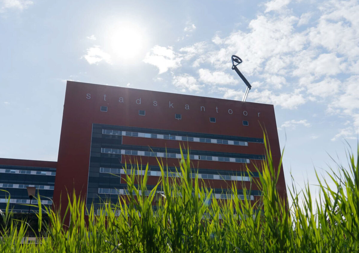Modern municipal office building of Environmental Authority IJsselland with a red facade and the lettering &laquo;Stadskantoor,&raquo; photographed from a low angle behind tall green grass under a sunny sky.