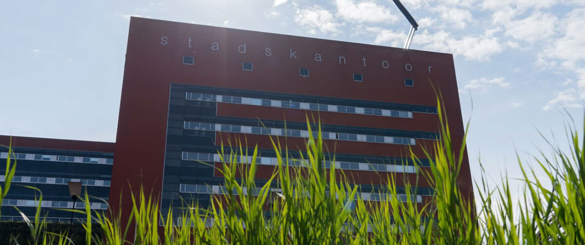 Modern kantoorgebouw van de Omgevingsdienst IJsselland met een rode gevel en de tekst 'Stadskantoor'. De foto is vanuit een laag standpunt genomen, waarbij hoog groen gras op de voorgrond zichtbaar is. Op de achtergrond staan het gebouw en enkele bomen onder een heldere, zonnige blauwe hemel.
