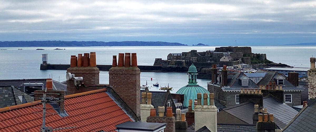 Panoramic view of a coastal town with historic rooftops, brick chimneys, and a green-domed building overlooking a calm harbor. A stone fortress extends into the sea under a cloudy sky, with distant shoreline visible on the horizon.
