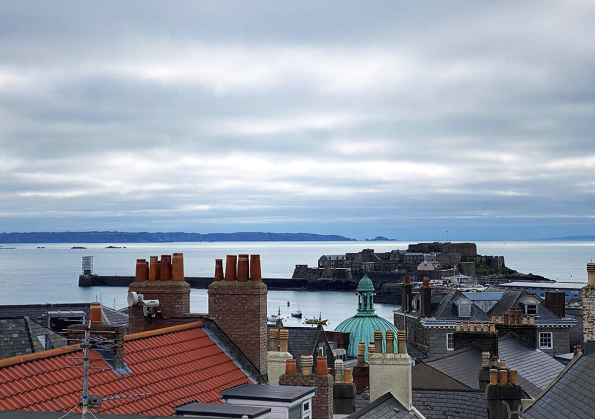 Panoramic view of a coastal town with historic rooftops, brick chimneys, and a green-domed building overlooking a calm harbor. A stone fortress extends into the sea under a cloudy sky, with distant shoreline visible on the horizon.