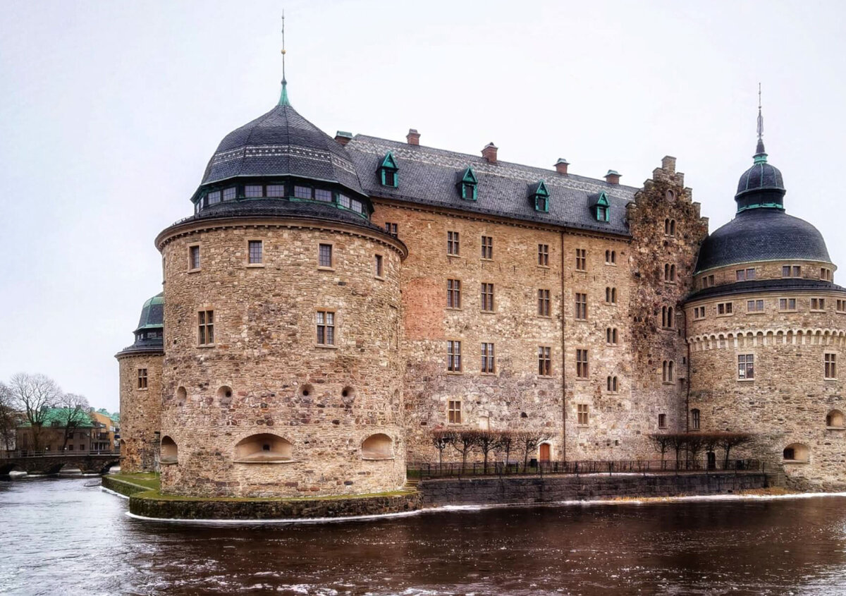 Historic &Ouml;rebro Castle in Sweden, a large stone fortress with round towers and dark domed roofs, surrounded by water under an overcast sky.