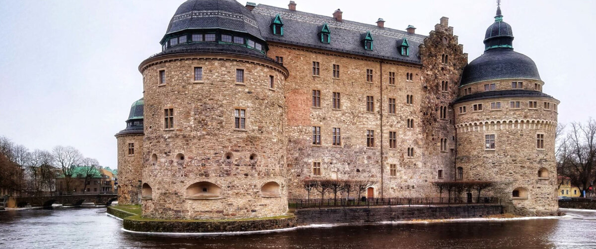 Historic &Ouml;rebro Castle in Sweden, a large stone fortress with round towers and dark domed roofs, surrounded by water under an overcast sky.