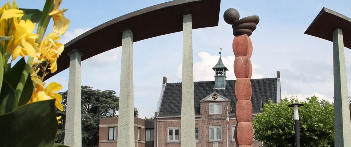 Town hall square in Someren, Netherlands, featuring a modern public art sculpture framed by curved stone pillars. Colorful flowers fill the foreground, while the municipal building and trees stand in the background under a clear blue sky.