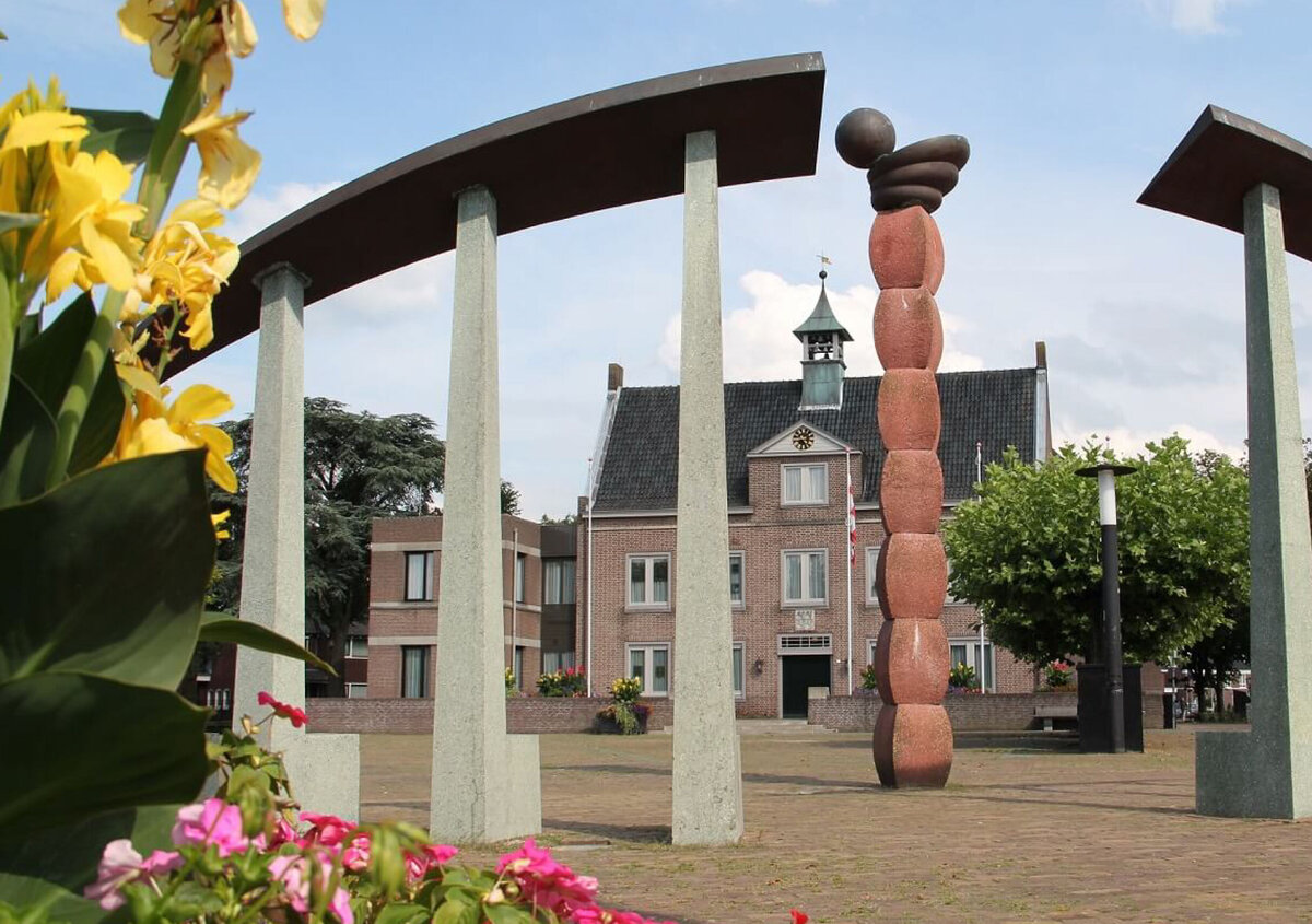 Town hall square in Someren, Netherlands, featuring a modern public art sculpture framed by curved stone pillars. Colorful flowers fill the foreground, while the municipal building and trees stand in the background under a clear blue sky.