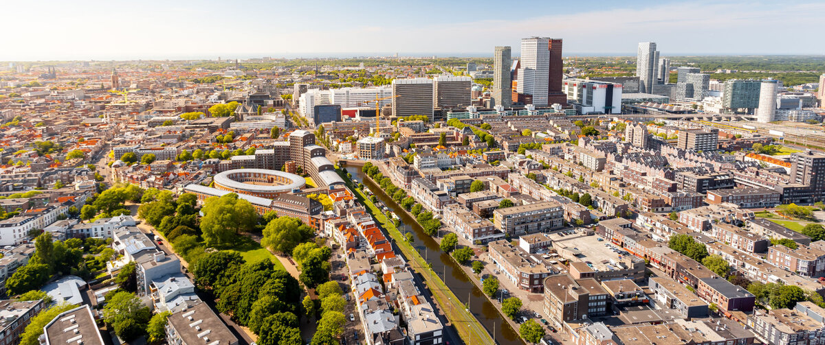 Aerial view of an expansive urban landscape on a sunny day. In the foreground, residential buildings, tree-lined streets, and a straight canal can be seen running through the neighborhood. In the center of the image are densely built city districts featuring a distinctive circular building complex. In the background, a modern skyline rises with several high-rise and office buildings. Above the city stretches a clear blue sky with only a few clouds.