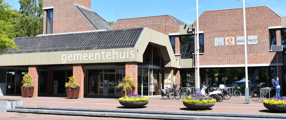 Frontvy av kommunhuset i Ermelo i Nederl&auml;nderna, med en tegelfasad, ett klocktorn och entr&eacute;n m&auml;rkt &raquo;gemeentehuis&raquo;. Framf&ouml;r byggnaden syns cyklar, blomsterplanteringar och flaggst&auml;nger under en klarbl&aring; himmel.