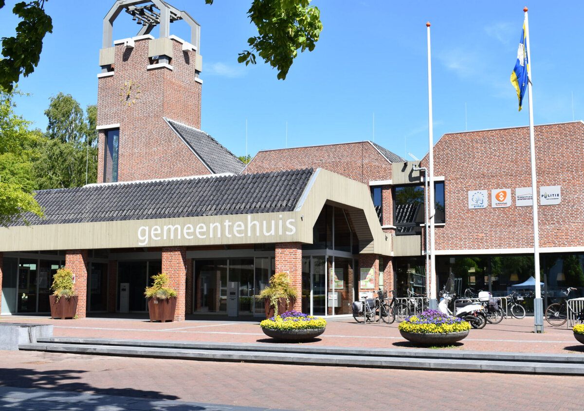 Front view of the Ermelo municipal building in the Netherlands, featuring a brick fa&ccedil;ade, a clock tower, and the entrance marked &laquo;gemeentehuis.&raquo; Bicycles, flower planters, and flagpoles are visible in front of the building under a clear blue sky.
