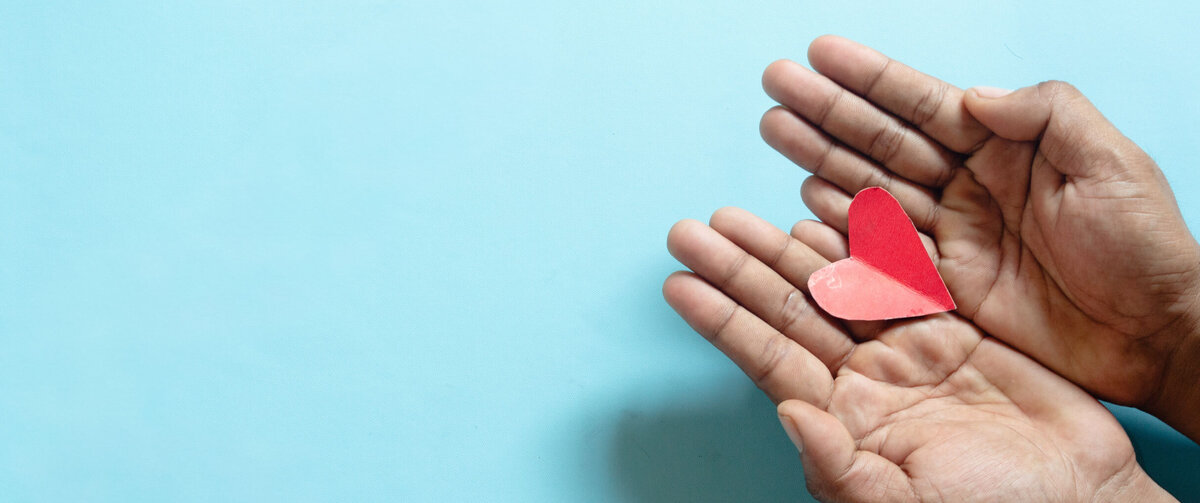 Two open hands gently hold a small red paper heart against a light blue background. The heart rests in the center of the palms and serves as the focal point of the image. The simple composition leaves ample empty space around the subject, while the soft colors convey themes of care, support, compassion, and social responsibility.
