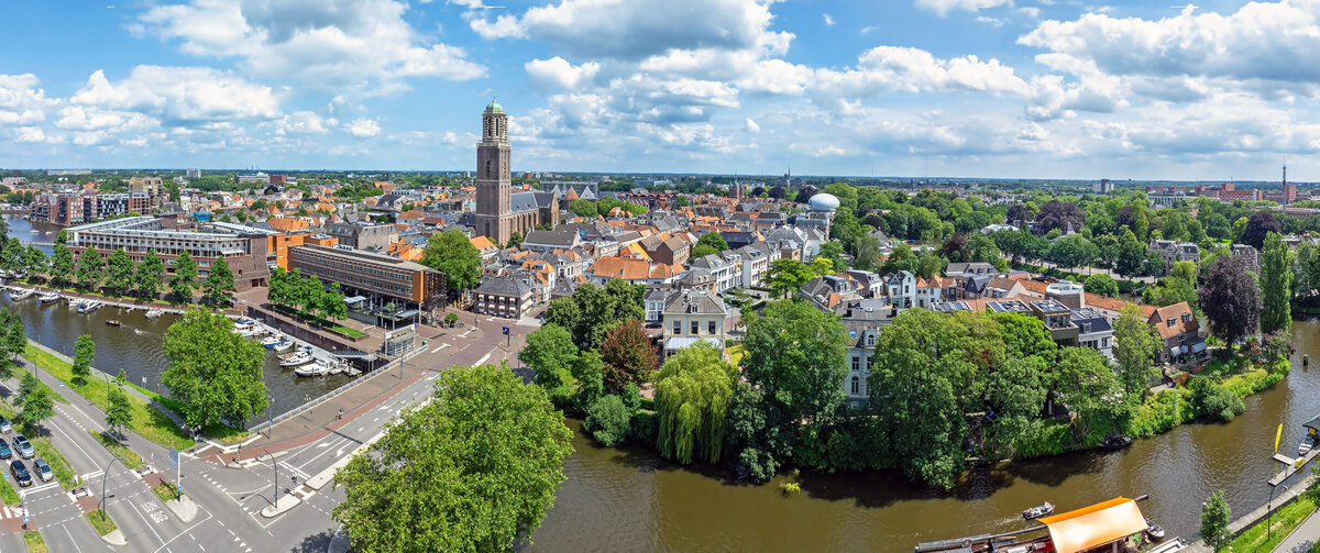 Aerial view of Zwolle's historic city center, featuring red-roofed buildings, a prominent church tower, canals, and a waterfront lined with boats. Dense urban streets and traditional architecture dominate the foreground, with residential neighborhoods and green areas extending into the background.