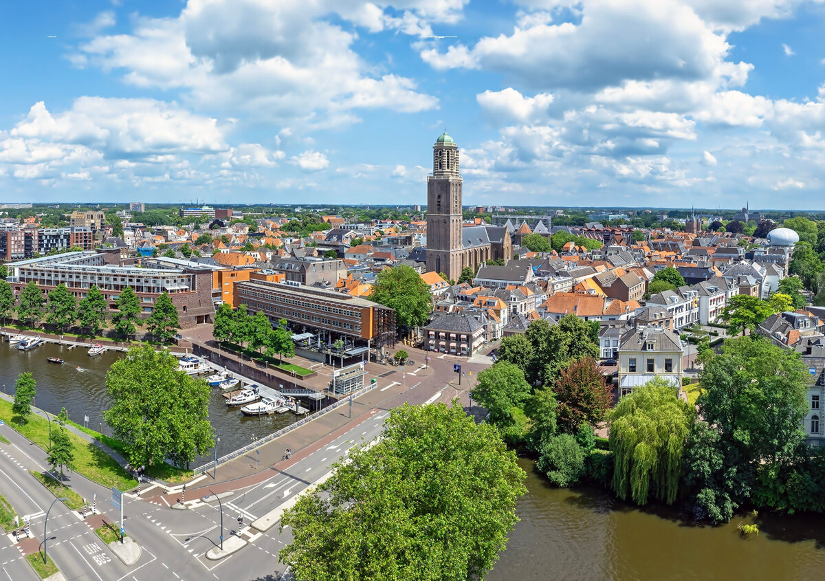 Aerial view of Zwolle's historic city center, featuring red-roofed buildings, a prominent church tower, canals, and a waterfront lined with boats. Dense urban streets and traditional architecture dominate the foreground, with residential neighborhoods and green areas extending into the background.