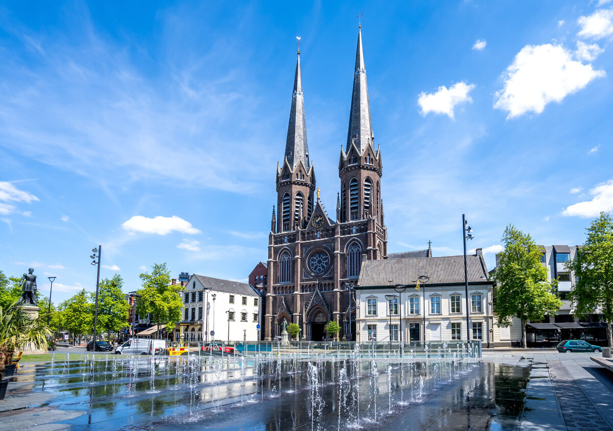 Kerk met twee torens die uitkijkt over een stadsplein met fonteinen, historische gebouwen en bomen onder een helderblauwe hemel.