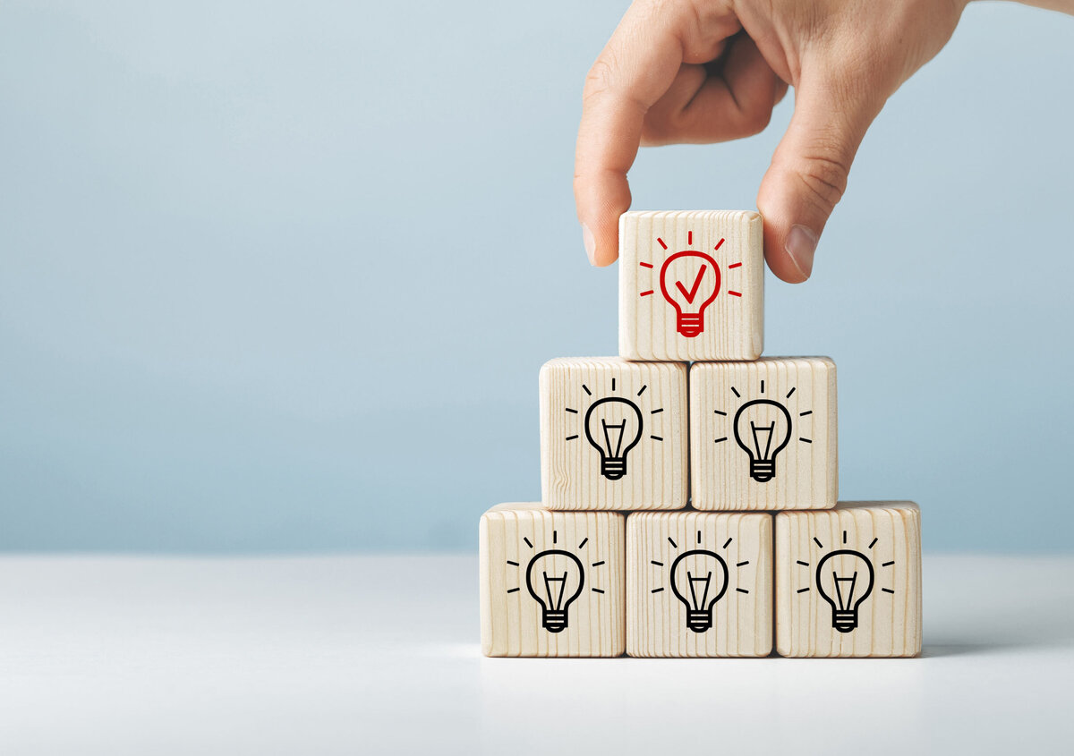 Hand placing a wooden block with a highlighted light bulb icon on top of a pyramid of blocks, symbolizing innovation, ideas, business solutions, and the discovery of new opportunities through a SmartDocuments demo.