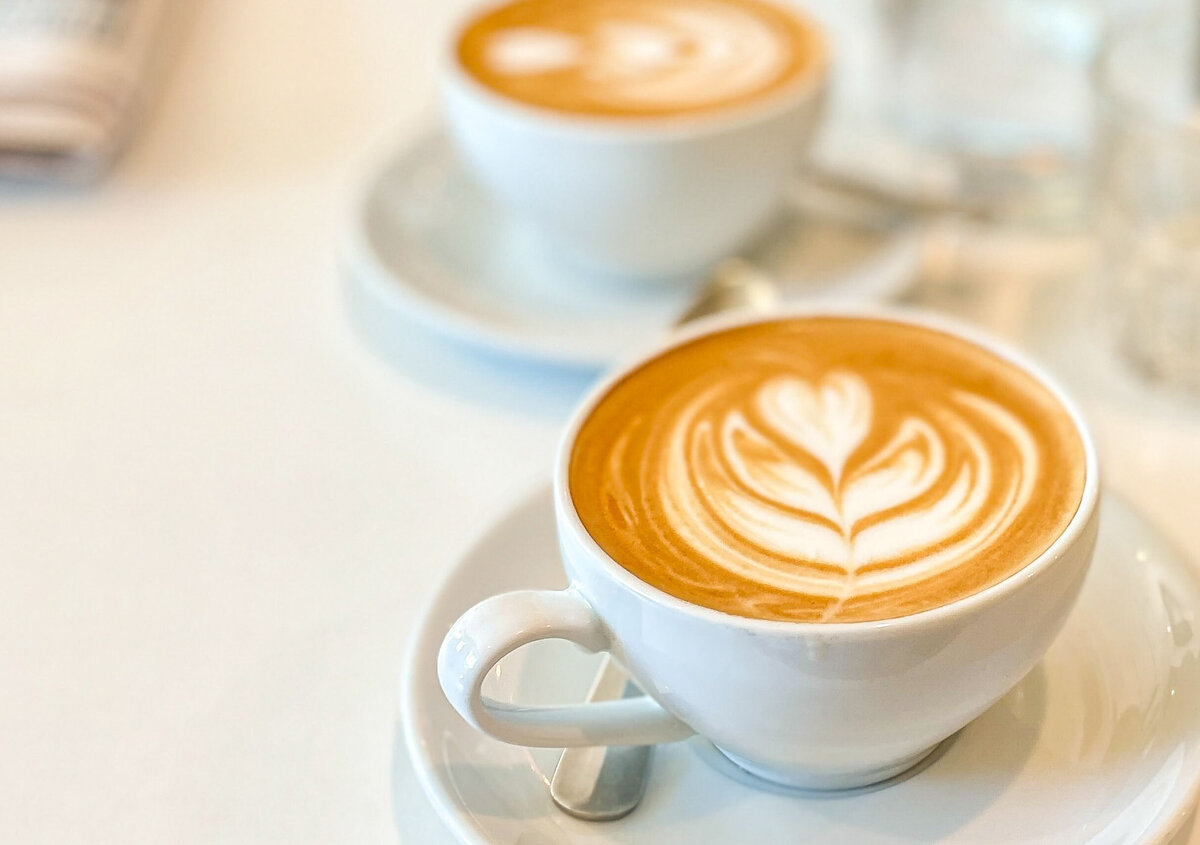 Zwei Tassen Cappuccino mit Milchschaumherz auf einem hellen Tisch. Die vordere Tasse ist scharf fokussiert, w&auml;hrend die hintere Tasse und der Hintergrund unscharf dargestellt sind. Daneben liegen eine Zeitung und ein Wasserglas.