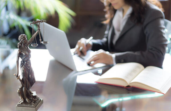 Fotografie eines Schreibtisches mit der Justiziafigur. Im Hintergrund ist eine Frau am Dargestellt mit Notebook.