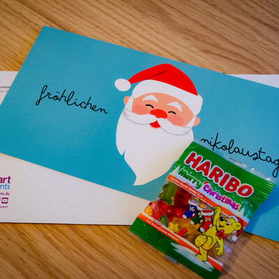 Bild vergrößern: Aufnahme Voderseite: Nikolausmailing für Teilnehmer:innen des Webinars "Zusatzmodule Serienbriefe + E-Mail-Plugin". Fotografie Schreibtisch mit Briefumschlag und Mailing sowie einer Tüte Haribo.